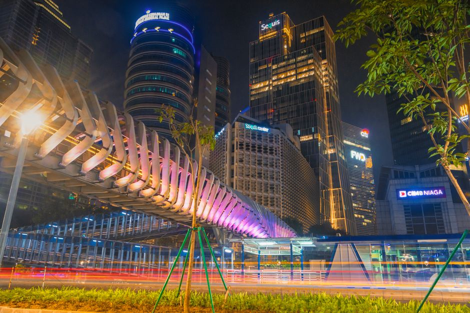 Dynamic night view of Jakarta's business district with illuminated skyscrapers and pedestrian bridge.
