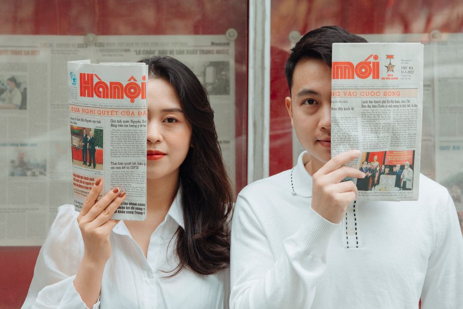 Asian couple playfully posing with Hanoi newspapers indoors, capturing love and connection.