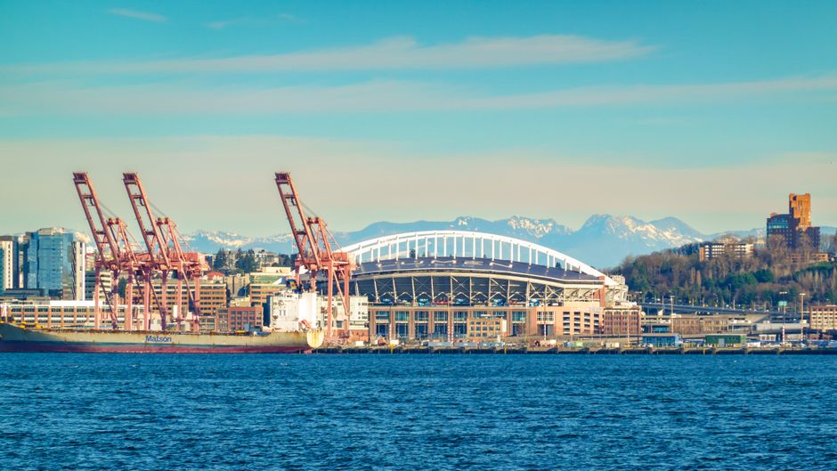 A scenic view of Seattle's waterfront featuring a stadium, cranes, and mountains.