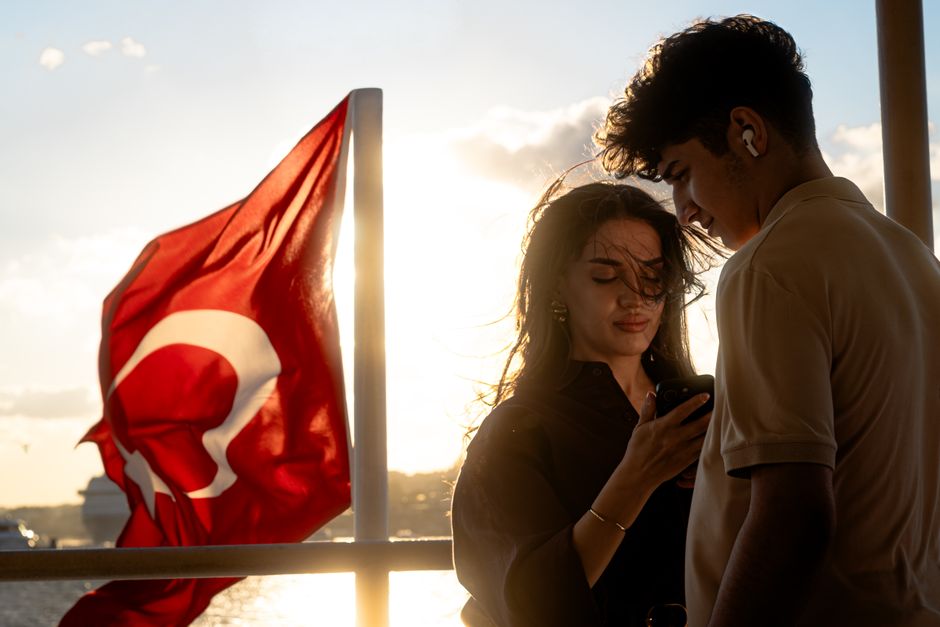 A couple enjoys a sunset cruise with a Turkish flag in the background, embracing summer romance.