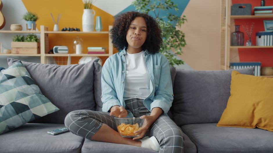 Young woman eating chips on a couch in a modern living room with colorful decor.