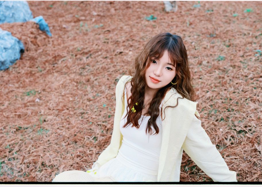 Young woman in white sitting on a bed of pine needles outdoors, smiling.
