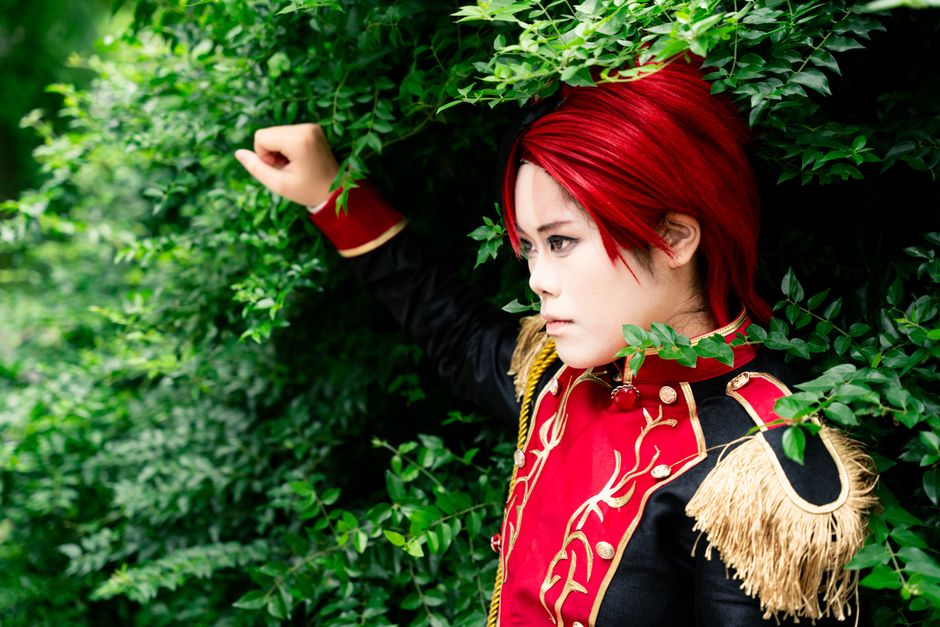 Cosplayer in intricate costume poses amidst vibrant green foliage, showcasing striking red hair and detailed attire.