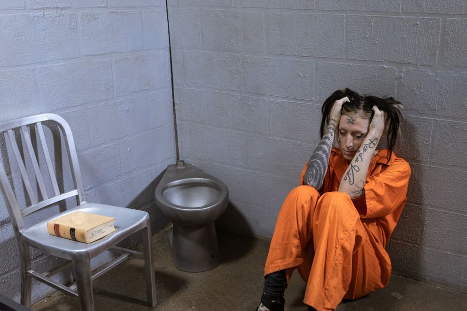 A female prisoner in an orange jumpsuit sits on the floor of a cell looking distressed, tattooed arms on head.