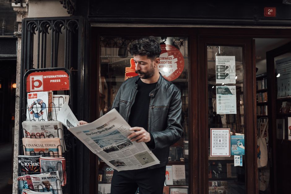 Serious bearded male with trendy haircut in casual clothes reading newspaper while standing near entrance of shop
