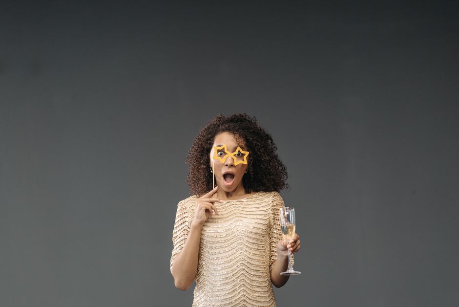 Astonished woman in gold outfit with star-shaped glasses, holding champagne glass against a plain background.