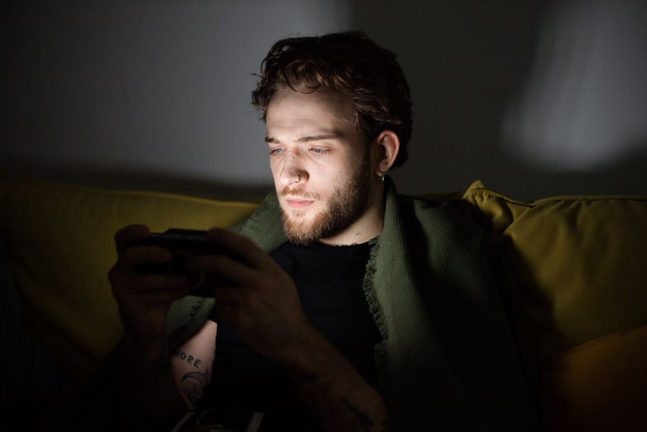 A bearded man playing video games indoors, focused on a handheld console.