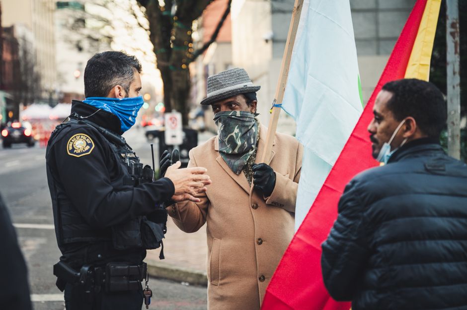 Side view of concentrated policeman in uniform standing and talking with African American men in protective mask and casual clothes on city street in daytime