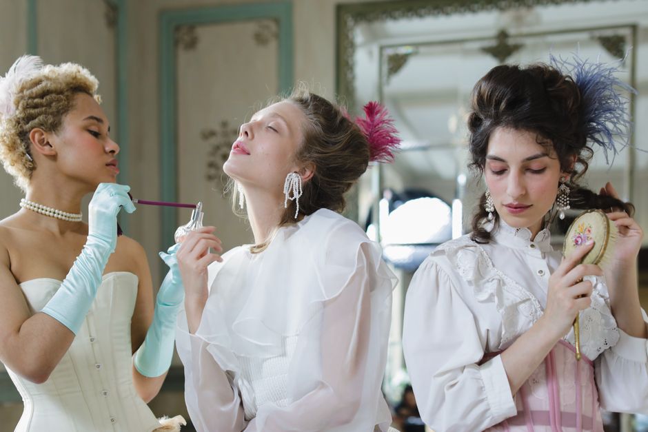 Three women in vintage period costumes pose elegantly indoors, capturing a sense of elegance and glamour.