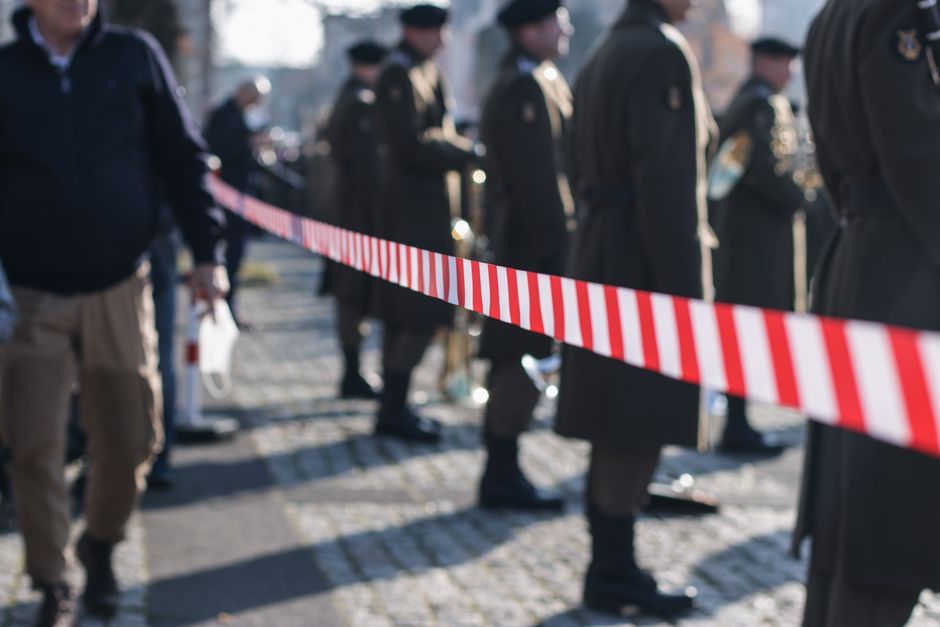 Soldiers in uniform stand behind a red-striped barricade during a military parade outdoors.