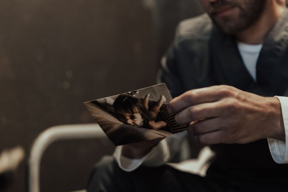 A man in prison uniform holds a photo of a child, evoking emotion and introspection.