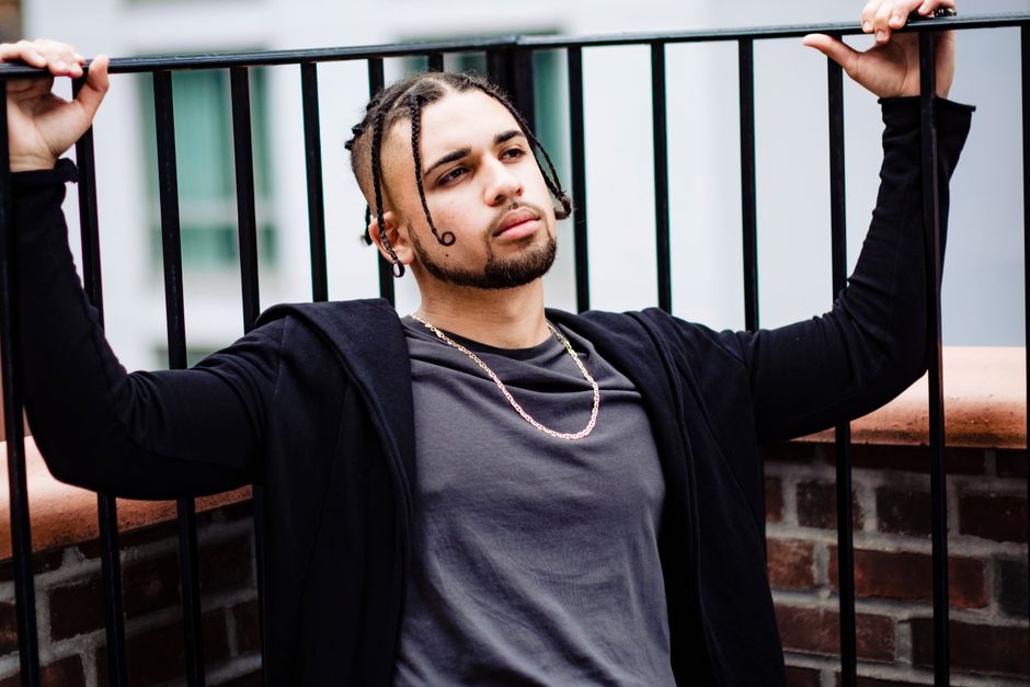 Portrait of a young man with braided hair and a stylish outfit leaning on a metal fence outdoors.