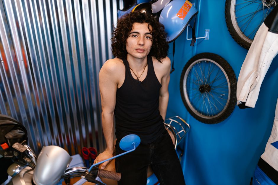 A young man in a black tank top leaning on a vintage motorbike in an indoor setting.