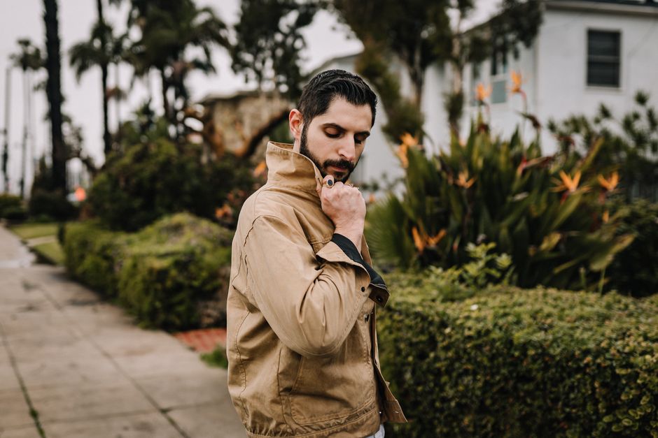 A fashionable man with facial hair adjusts his brown jacket outdoors.