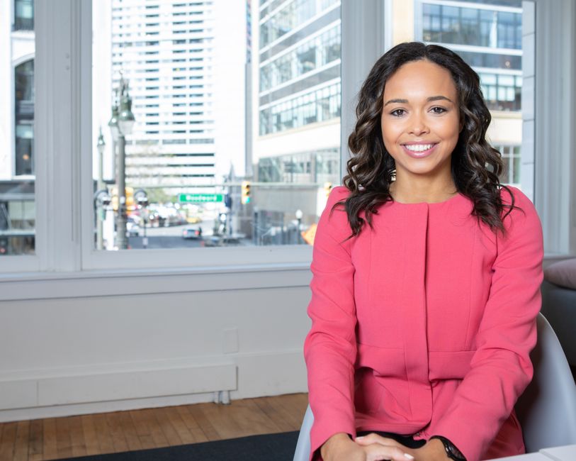 Young woman in a pink blazer smiles confidently in a modern city office setting.