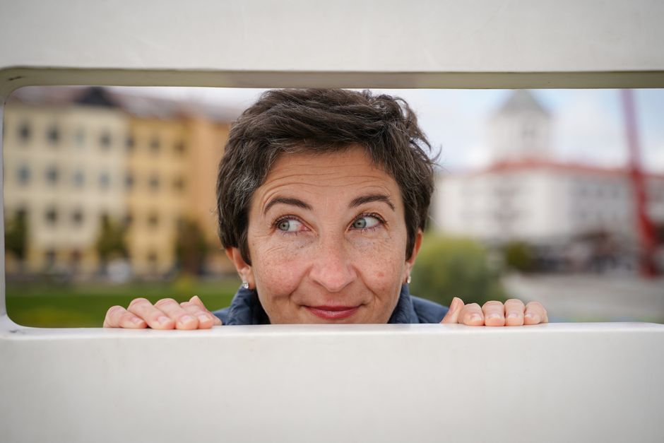 A cheerful woman peeks through a bench in Jelgava, Latvia, with buildings in the background.