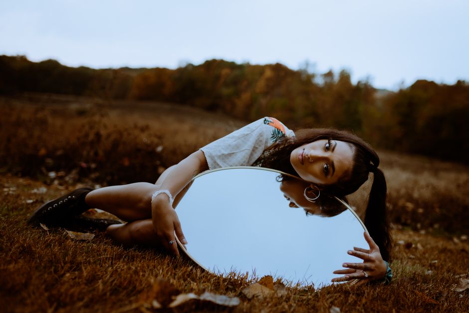 Artistic portrait of a woman lying on grass with a round mirror reflecting the sky during fall.