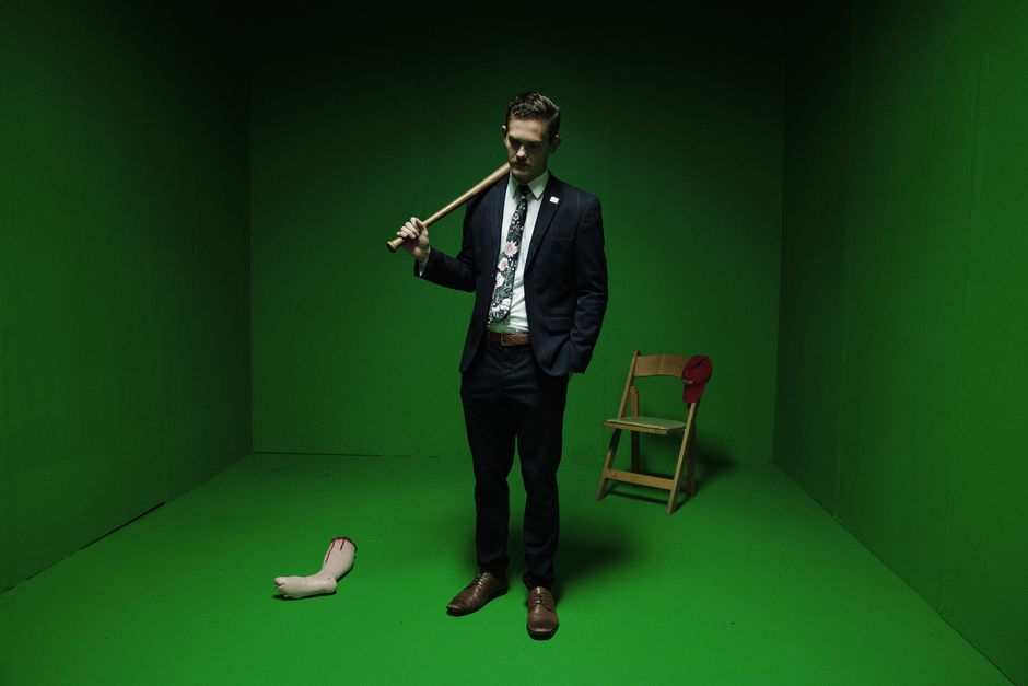 A man in a suit stands in a green room, holding a baseball bat over his shoulder.