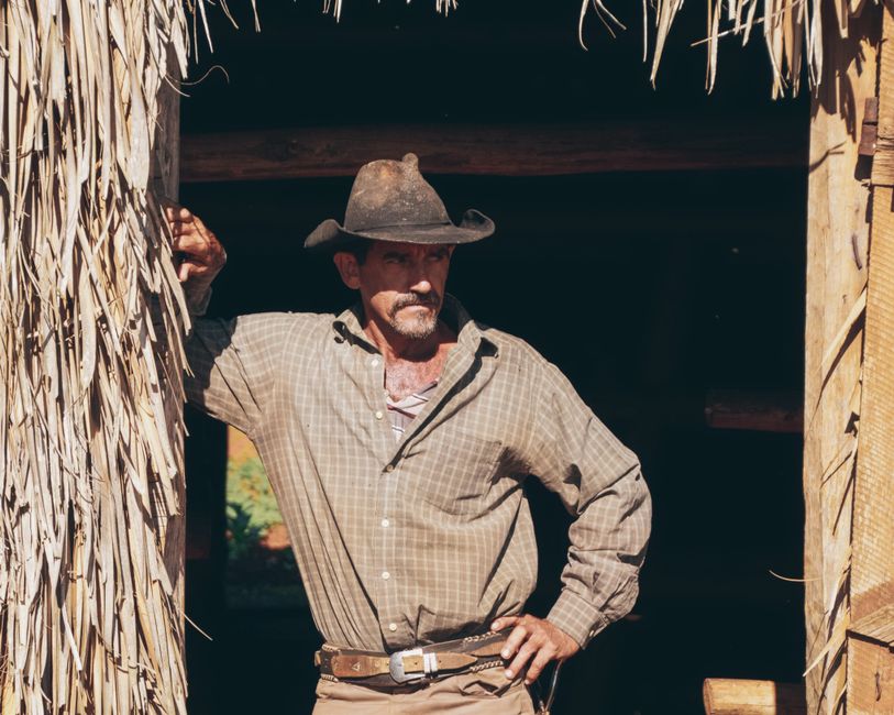 A senior cowboy stands confidently in a sunlit barn doorway, wearing a hat and rugged shirt.