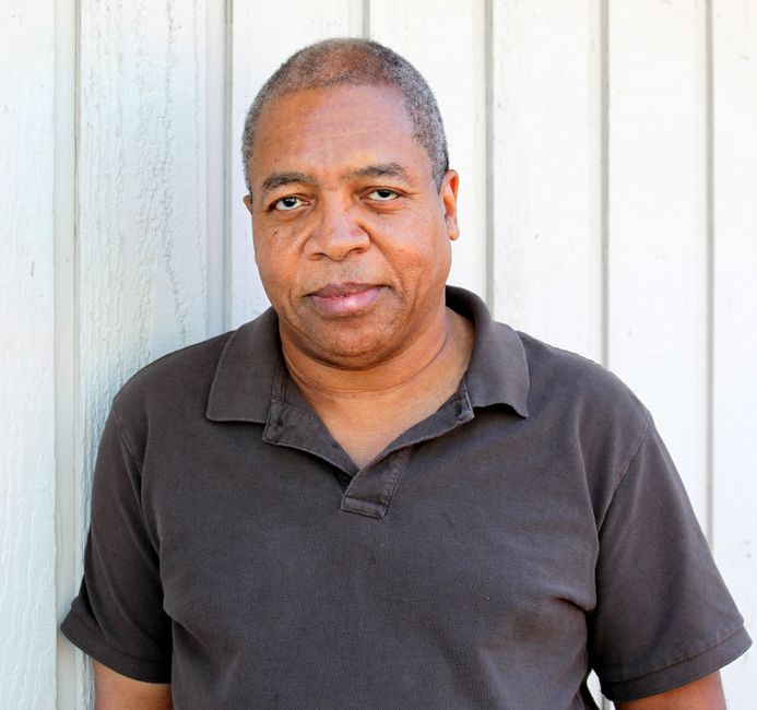 Casual portrait of a mature man standing against a light wall wearing a dark polo shirt.