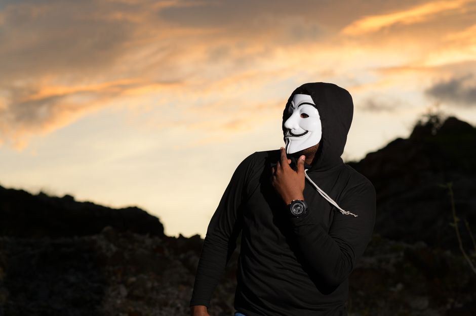 Man wearing a Guy Fawkes mask standing outdoors at sunset, creating a mysterious silhouette.