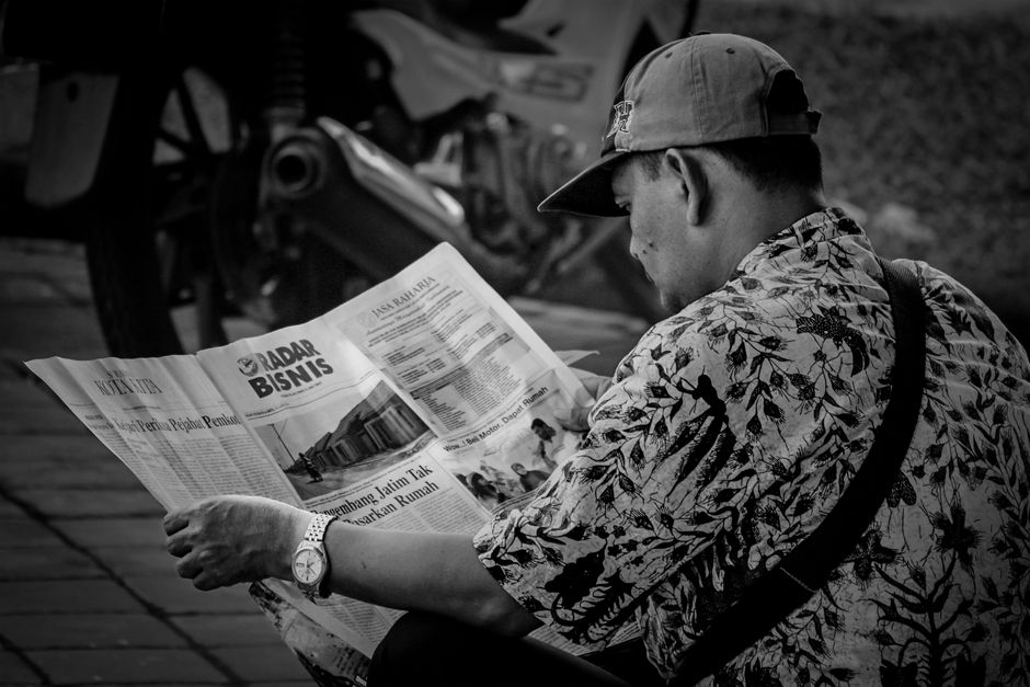 Black and white photo of a man reading a newspaper outdoors in Surabaya, Indonesia.
