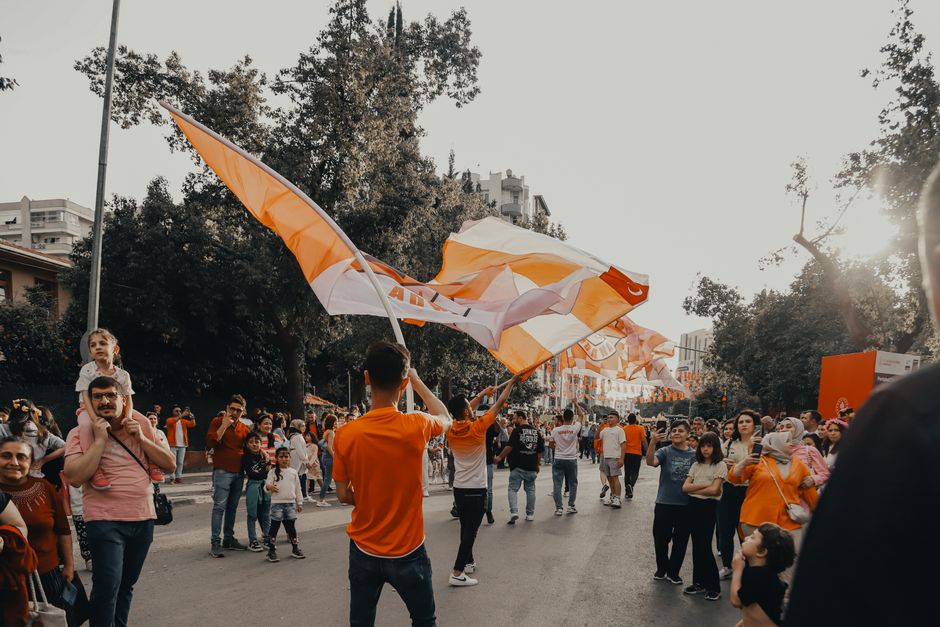 A lively city street parade with people celebrating under colorful flags and banners during the day.
