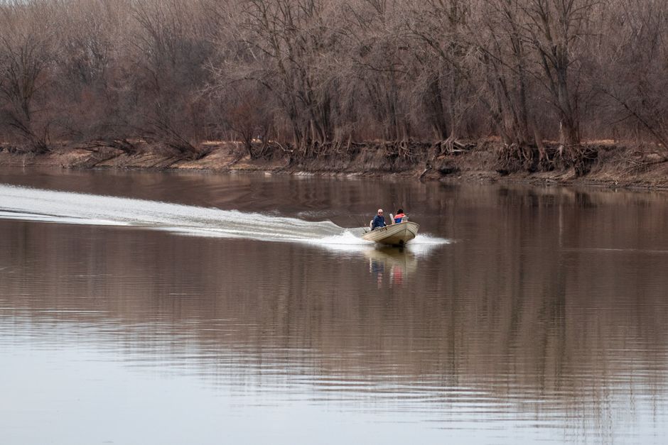 A small motorboat carrying two passengers glides through a calm river surrounded by leafless trees.