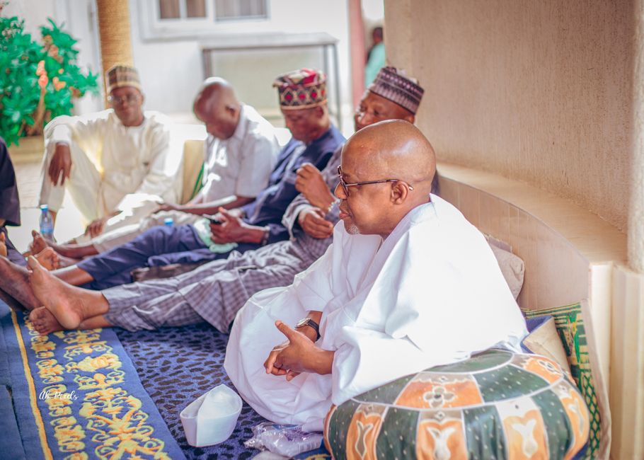 A group of African men sit indoors on a decorative mat, engaged in conversation.