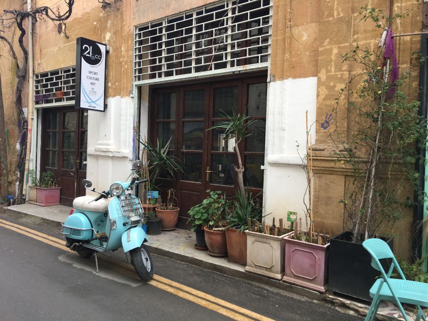 A charming turquoise scooter parked outside a rustic cafe in Nicosia, Cyprus, with potted plants and vintage decor.