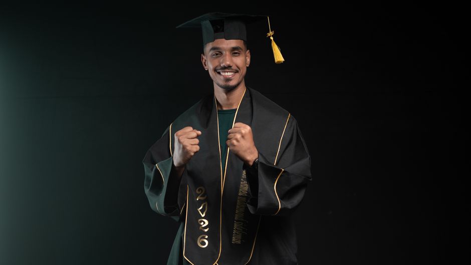 Excited young graduate in cap and gown celebrating success, fist raised, on a dark background.