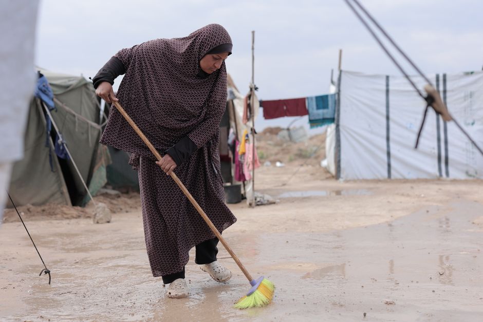Woman sweeping muddy ground in a refugee camp setting.