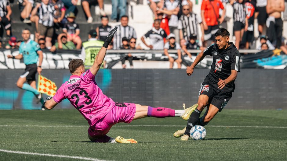 Goalkeeper in pink slides to block forward in black during a soccer match.