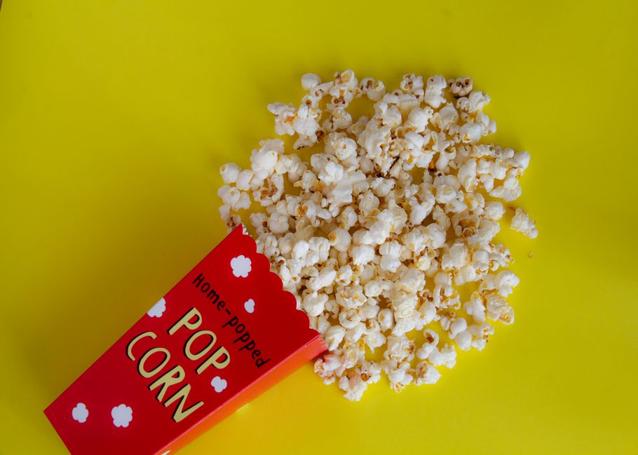 Top view of bright red decorative takeaway bucker with pile of crunchy popcorn spilled on yellow table