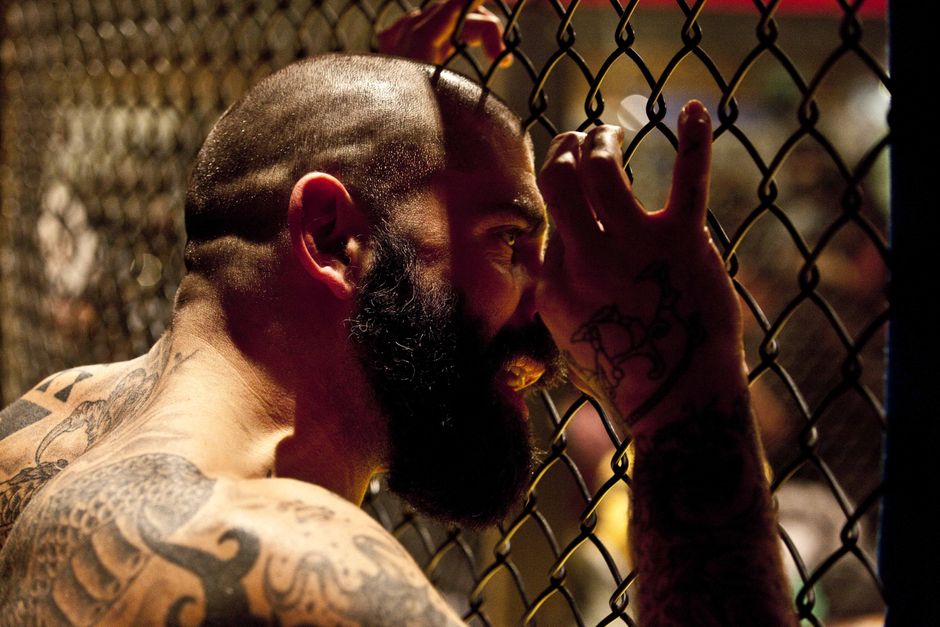 A bearded, tattooed man grips a chain-link fence, conveying intensity and solitude.
