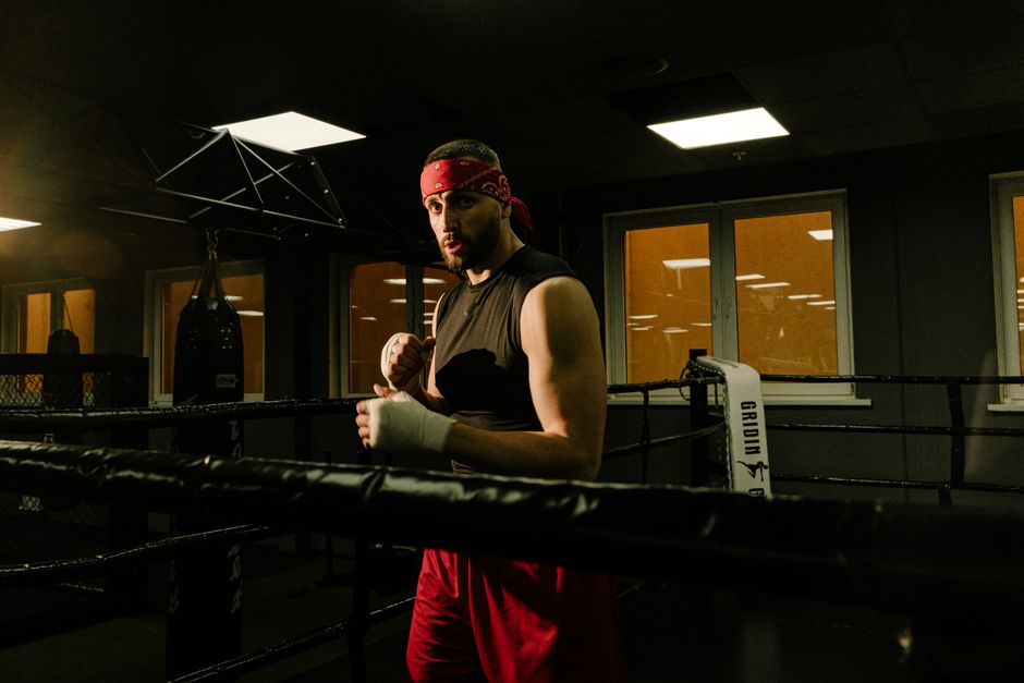 Man in boxing ring wearing red trunks and headband, preparing for a fight indoors.