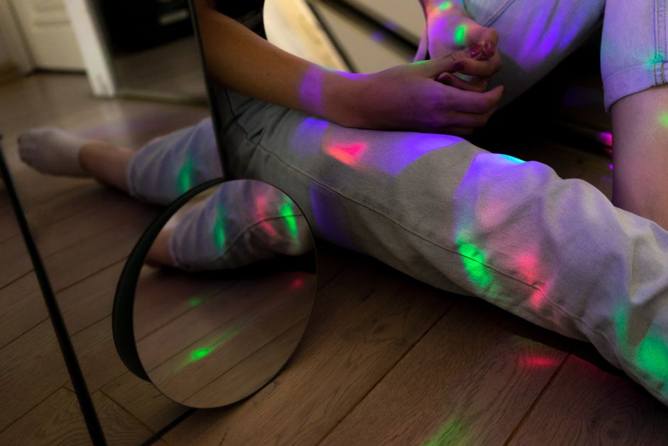 Crop unrecognizable person touching hands and sitting on floor surrounded by various mirrors in room with colorful lights