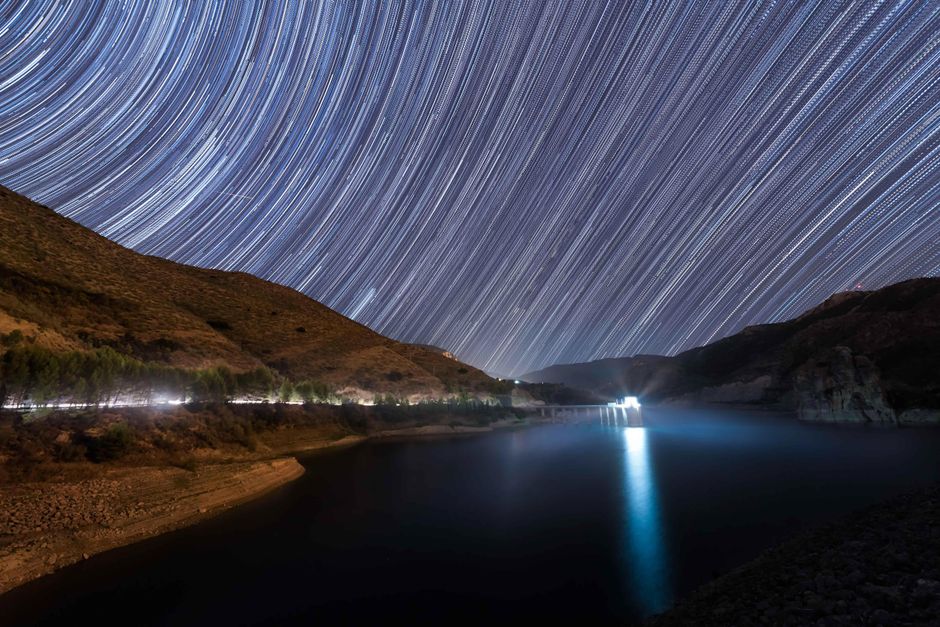 A mesmerizing long exposure capturing star trails over a serene lake and hills in Spain.