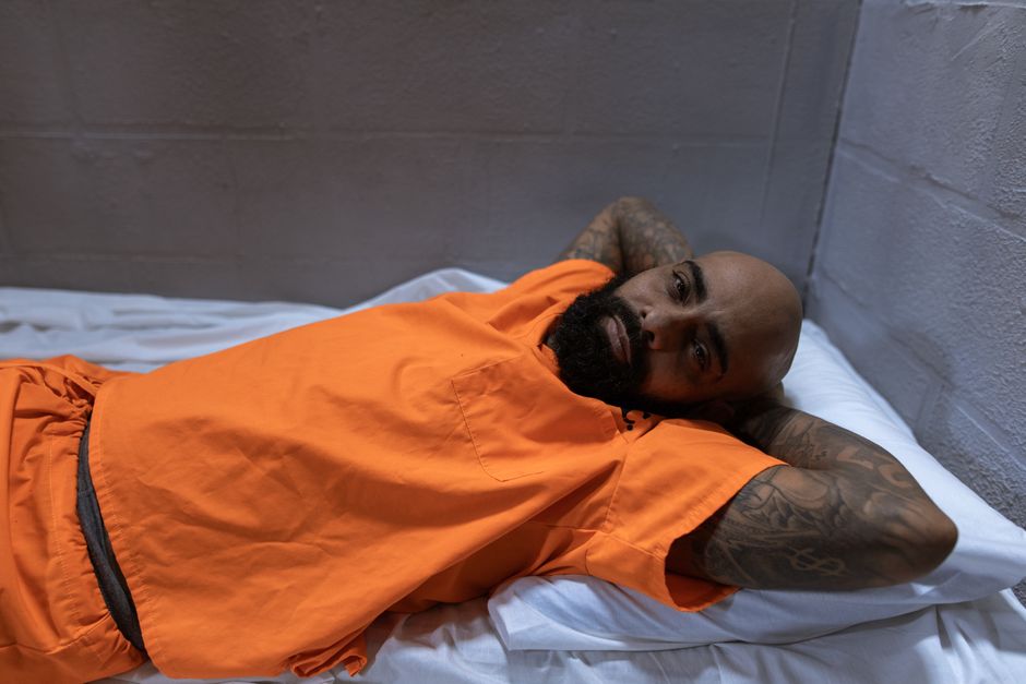 Man in orange prison uniform lying on bed, showing tattoos, indoors setting.