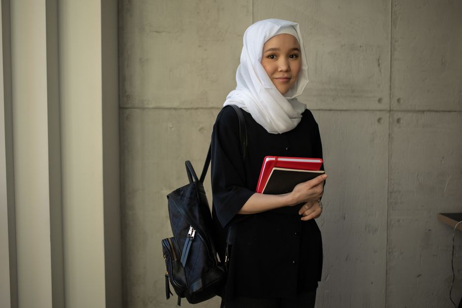 Cheerful Muslim female student in traditional headscarf with backpack looking at camera while standing near wall with notepads during studies in university