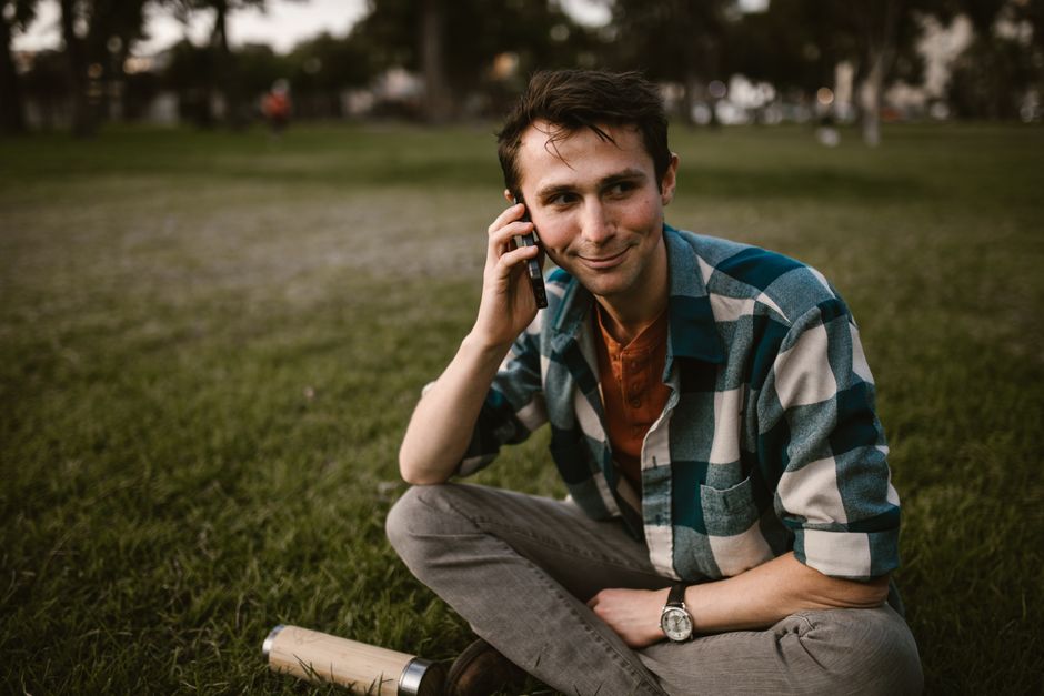A young man in plaid shirt and gray pants happily converses on phone while sitting on a park lawn.