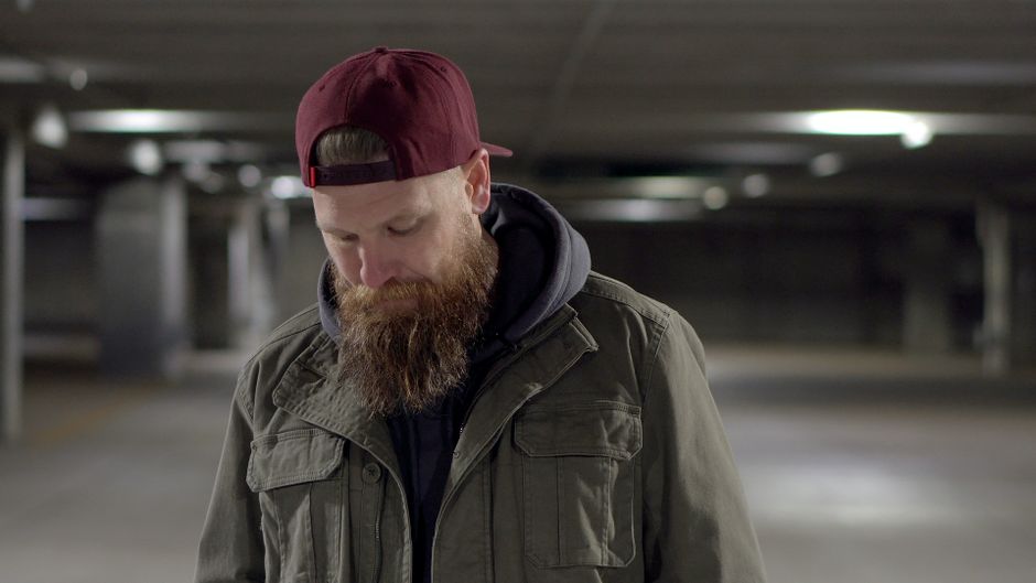 Casually dressed man with a beard in a semi-lit urban environment wearing a cap.