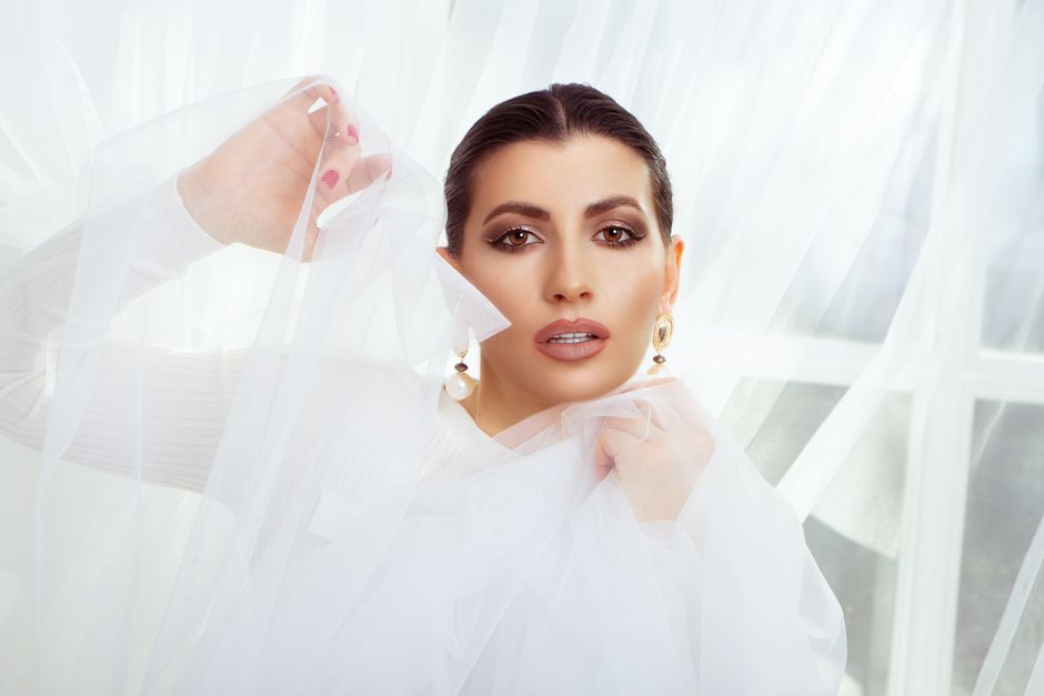Charming female with black hair and earrings looking at camera while standing with translucent white tulle near window in room