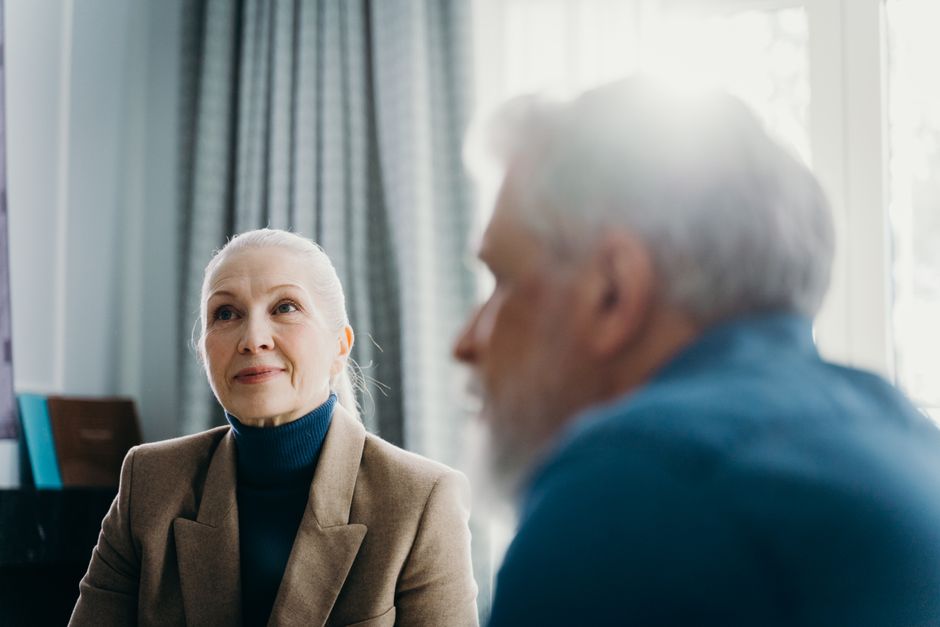 Elderly man and woman engaged in a warm conversation, sunlight streaming through the window.