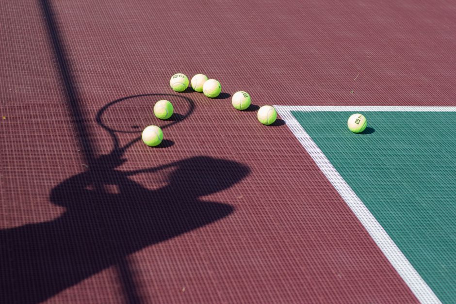 A tennis racket shadow and scattered balls on a colorful court under sunlight.