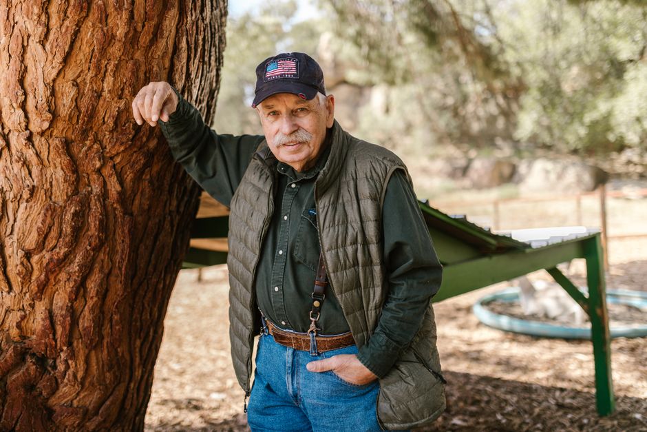 An elderly farmer with a warm smile leaning against a tree in a rural outdoor setting.