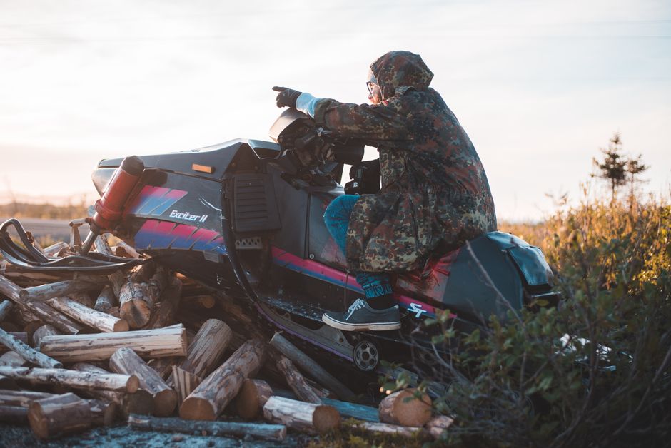 A man wearing a camouflage jacket points while sitting on a snowmobile atop a pile of logs outdoors.
