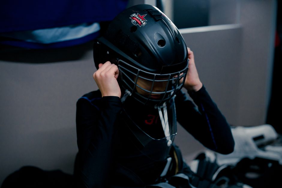 A teenage ice hockey goalie adjusts his helmet in a dressing room, preparing for a game.
