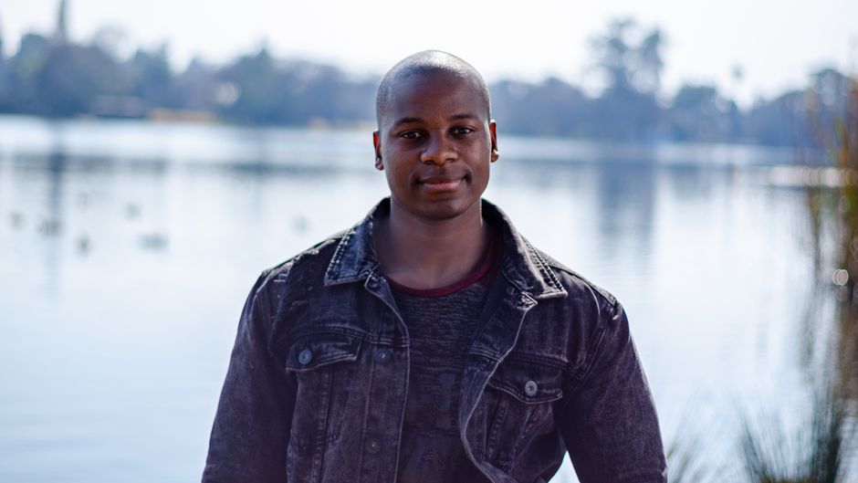 A smiling man stands outdoors by a serene lake, wearing a denim jacket.