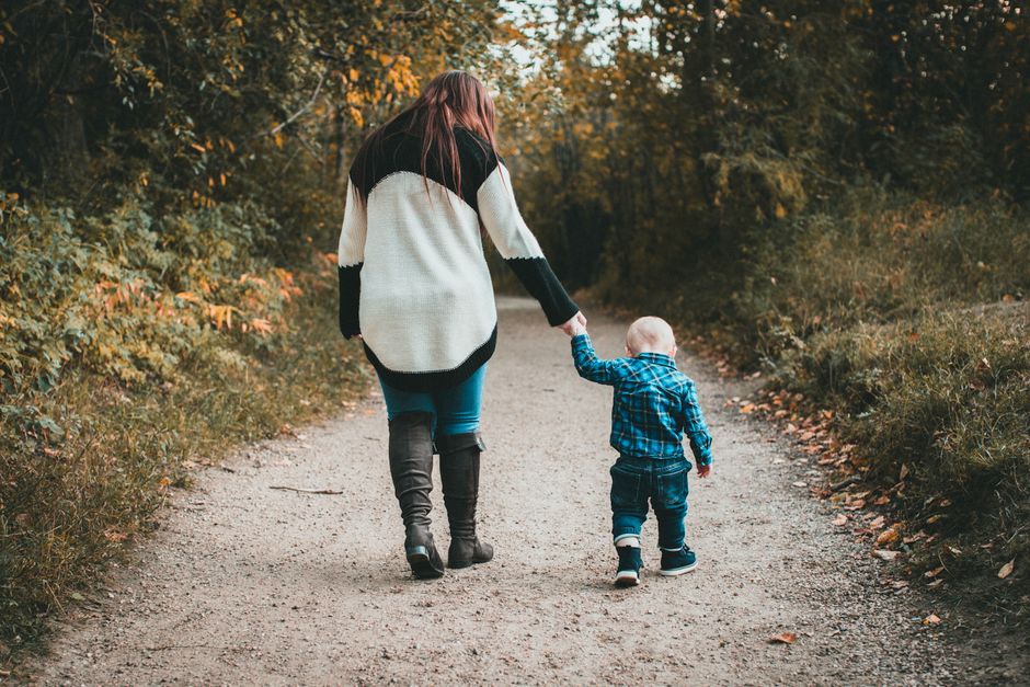 A mother and her son walk hand in hand on a forest path surrounded by autumn foliage.